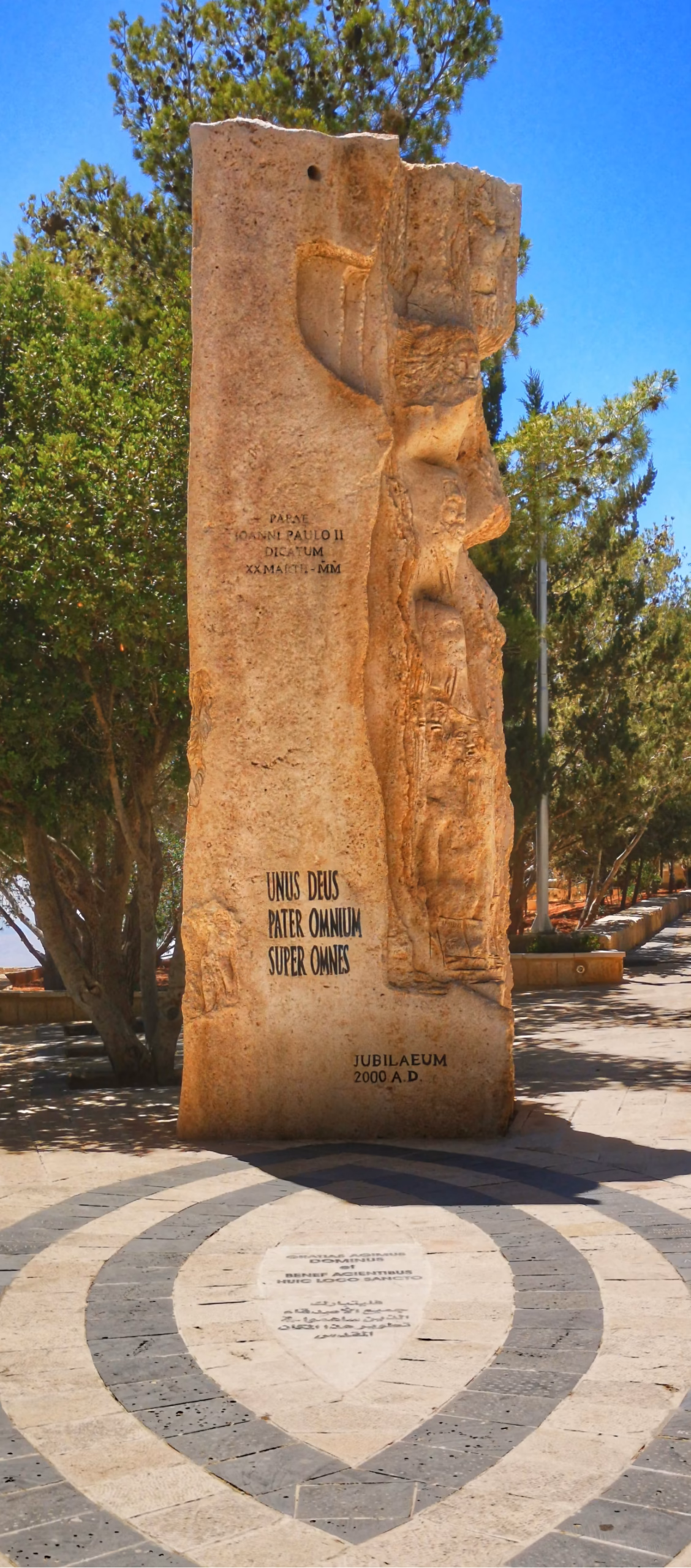 Tall inscribed stone stele on Mount Nebo, Jordan, bearing a relief carving and the Latin inscription Unus Deus Pater Omnium Super Omnes, surrounded by trees
