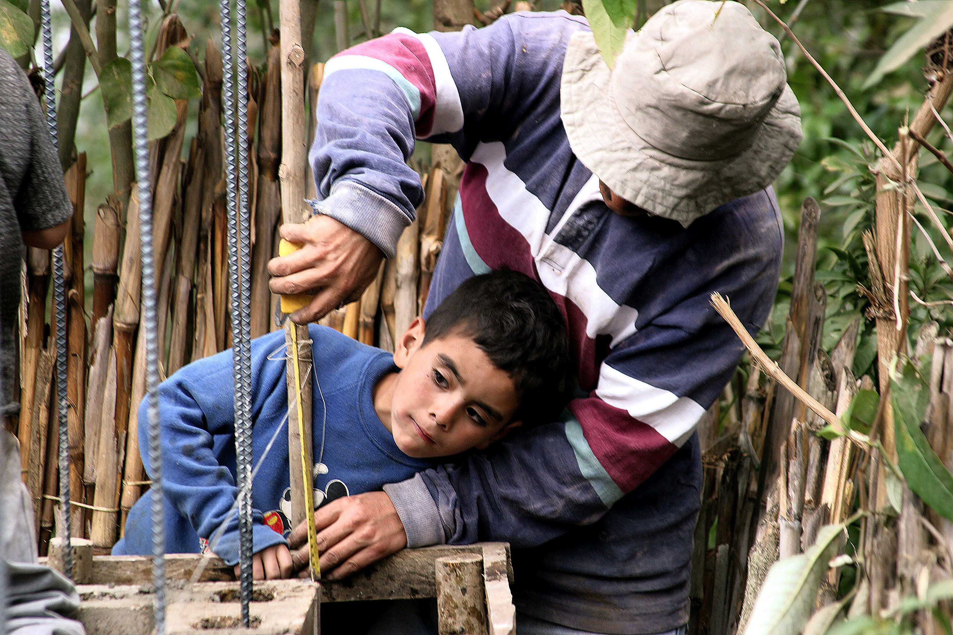 A man in a sun hat guides a boy's hands as they measure and lay reinforced concrete blockwork on a self-build house in Guatemala