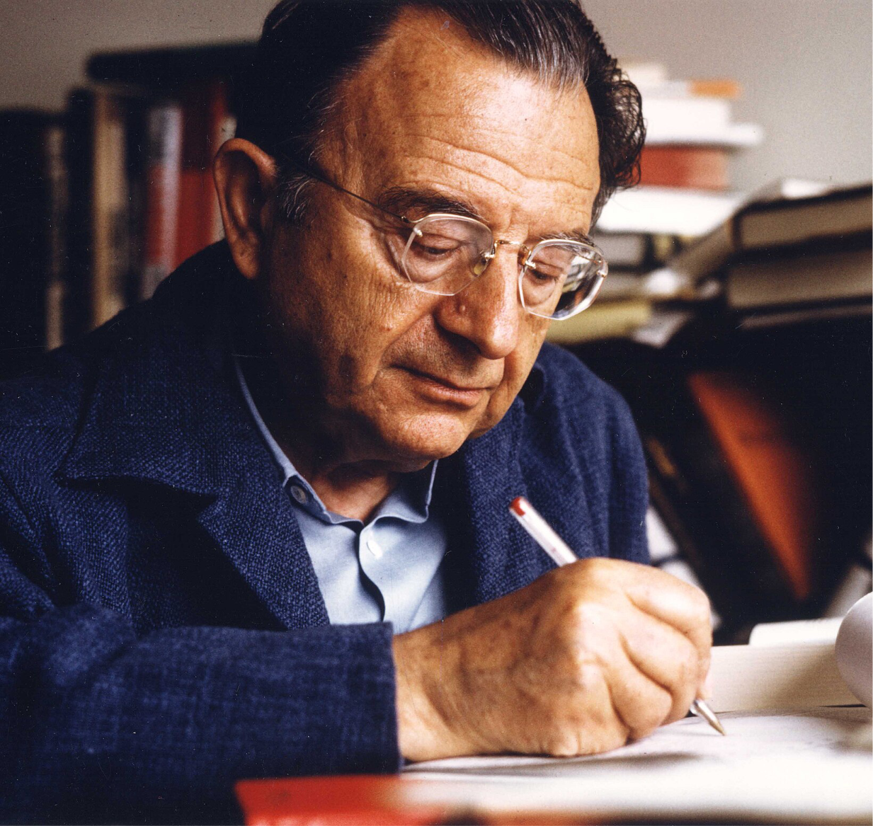 Colour photograph of Erich Fromm, wearing glasses, writing at a desk with books stacked behind him